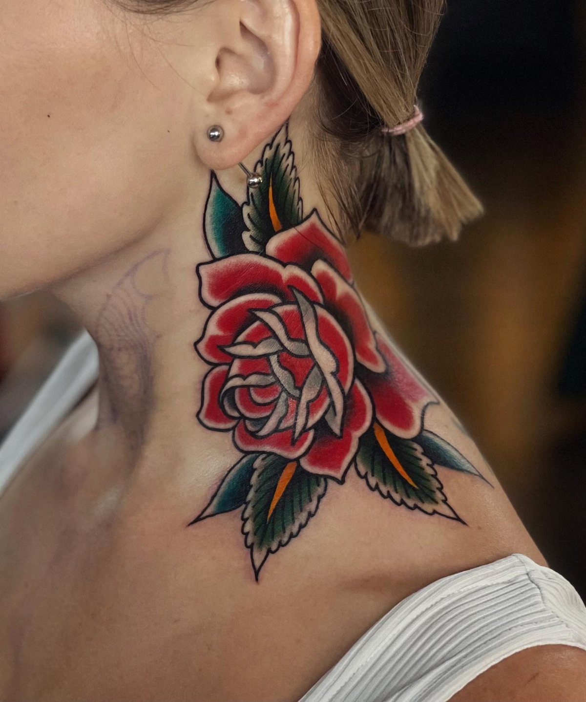 Traditional red rose tattoo with green leaves on a person's side neck.