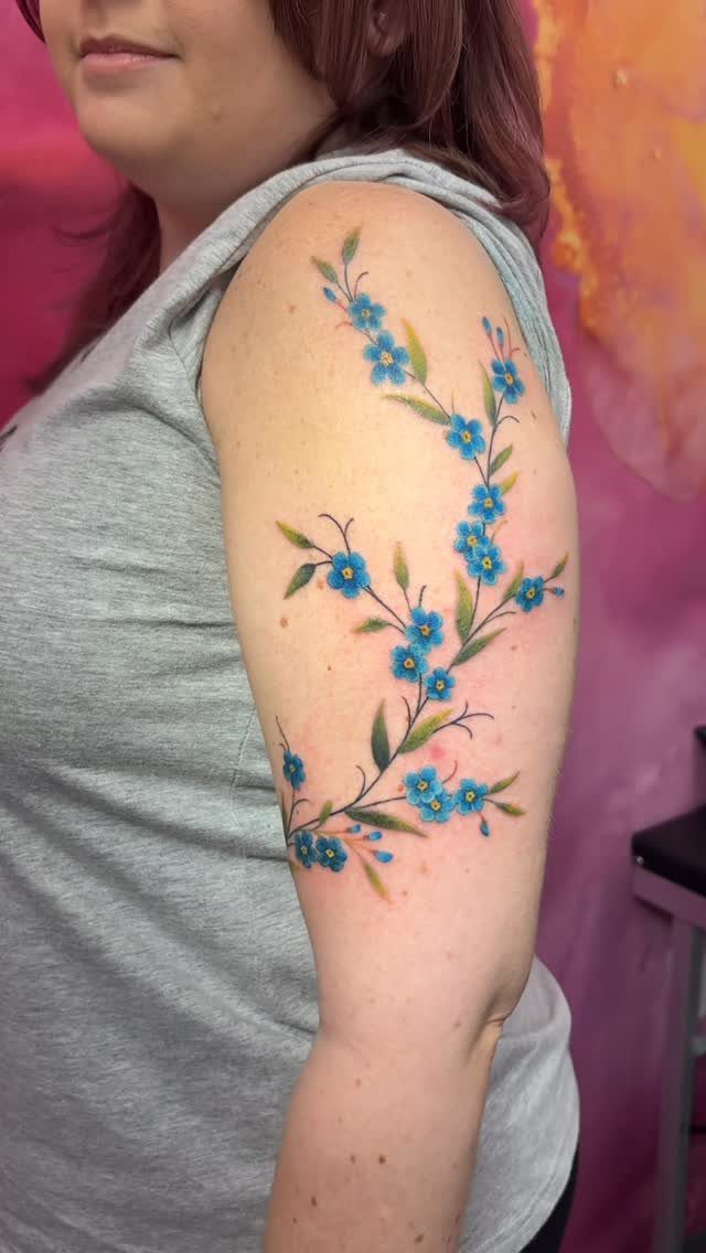 Blue forget-me-not flower tattoo with green leaves on a woman's upper arm.