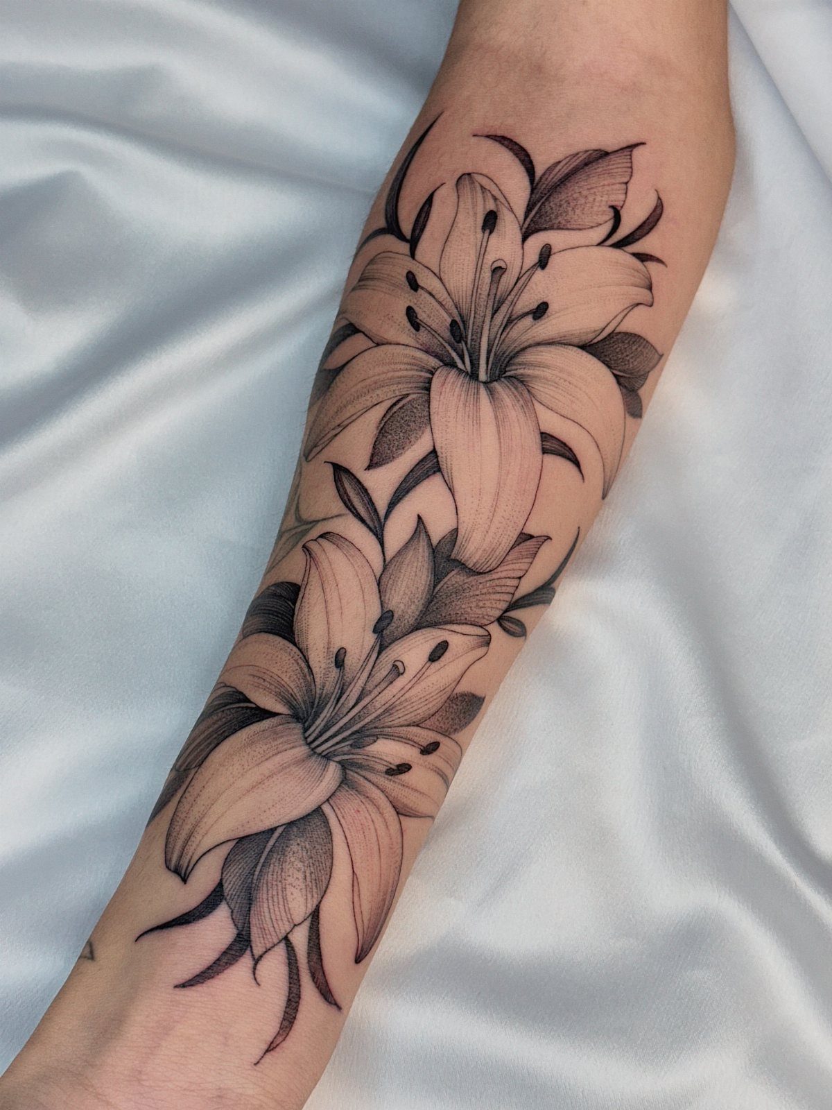 Two detailed lily flower tattoos in black and grey ink on a forearm.