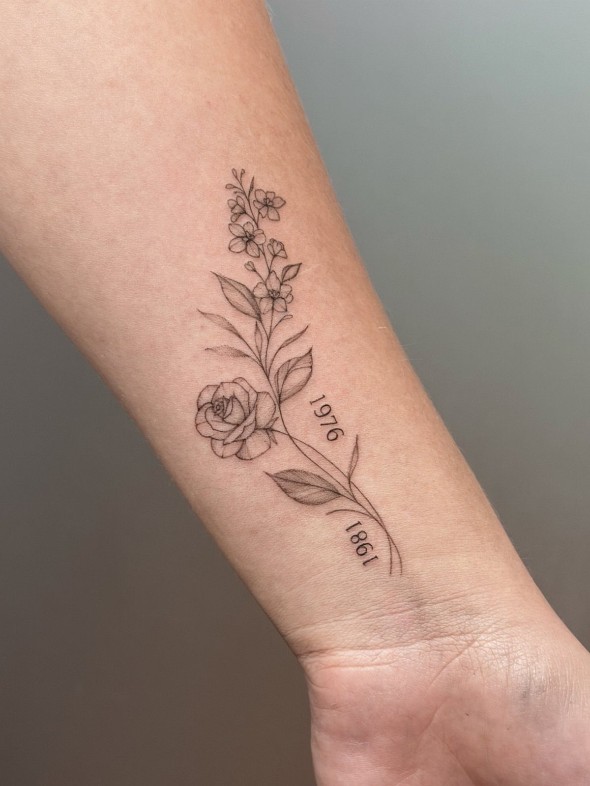 Fine-line floral tattoo on a forearm with birth flowers and years 1976 and 1981.