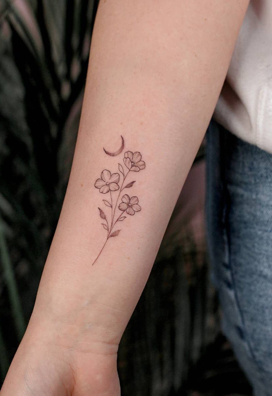Fine-line flower tattoo with a crescent moon on a person's forearm.