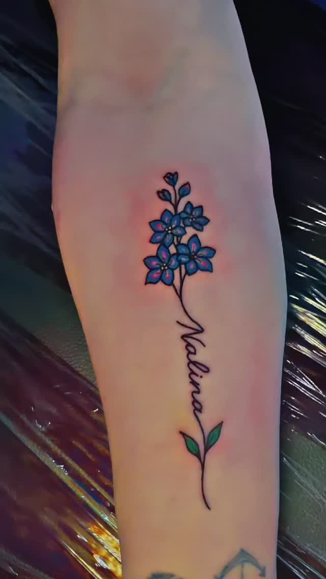 Blue flower tattoo on a forearm with the name Nalina written in cursive.