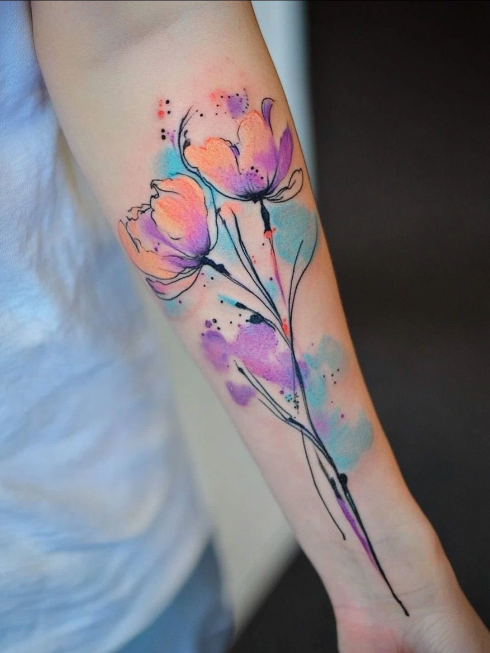 Watercolor flower tattoo on a forearm with purple and orange petals and blue splatters