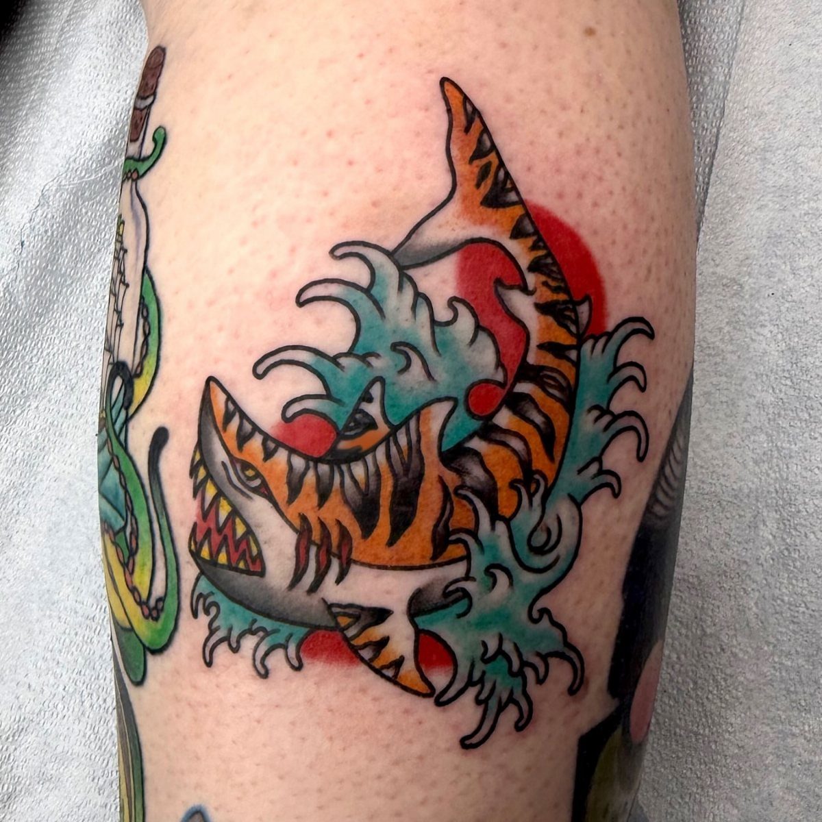 Tiger shark tattoo with orange stripes leaping through teal ocean waves and red sun.