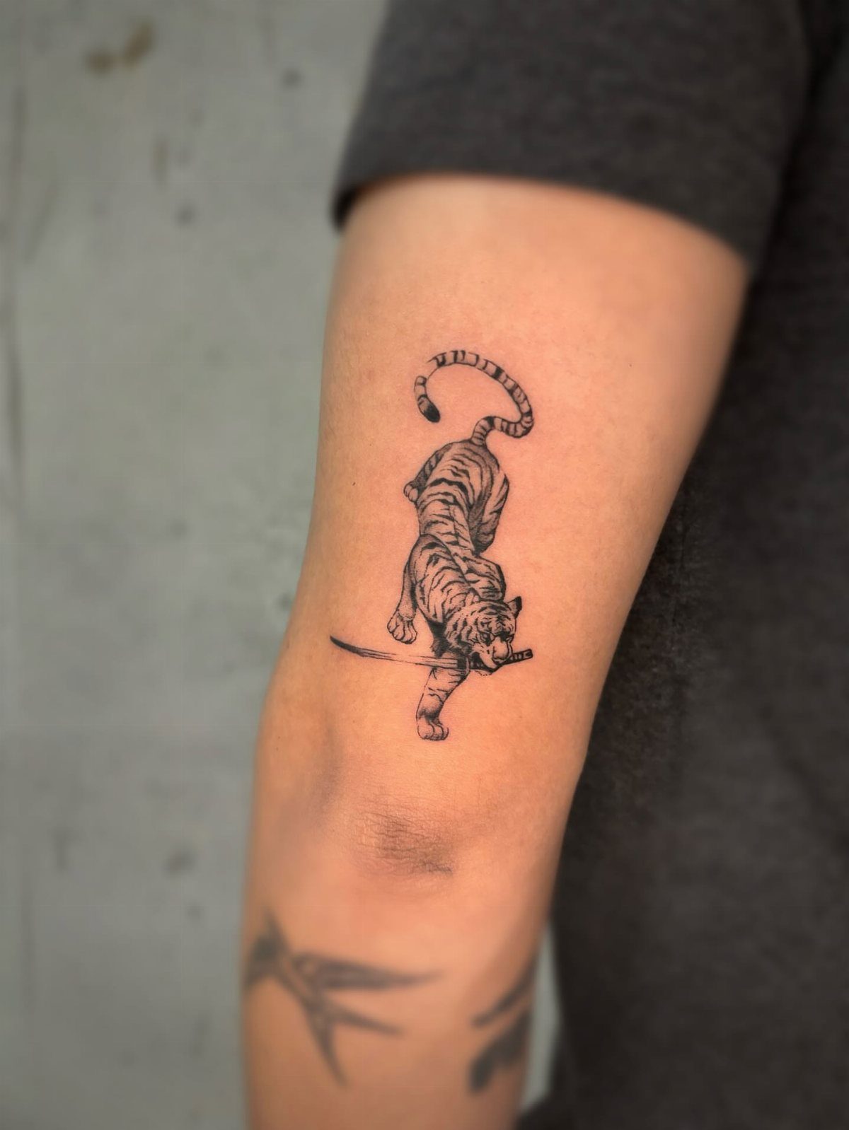 Small black ink tiger tattoo holding a sword on a person's upper arm.