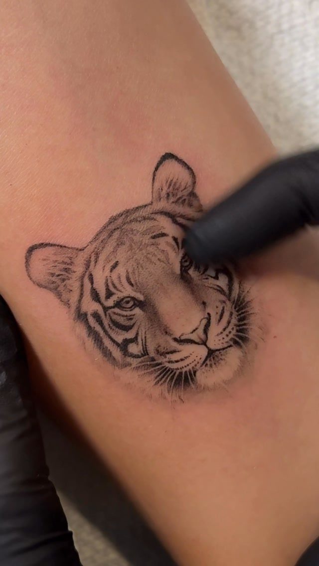 Tiger face tattoo in black and grey ink on a person's skin.