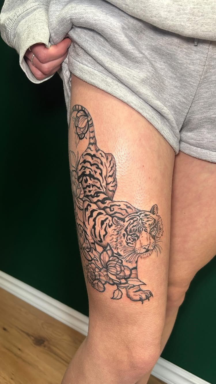 Tiger and flower tattoo on a person's upper thigh in black and grey ink.