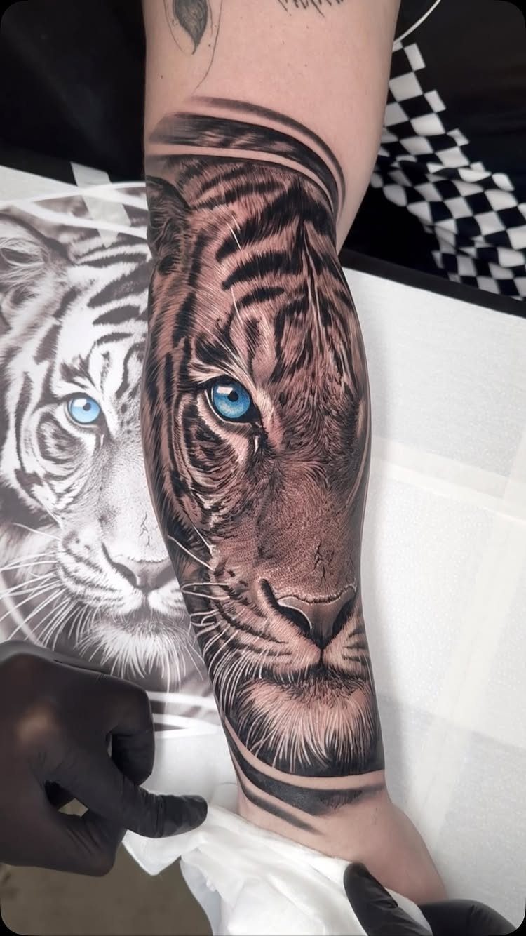Tiger face tattoo on a forearm with a vivid blue eye and detailed shading.