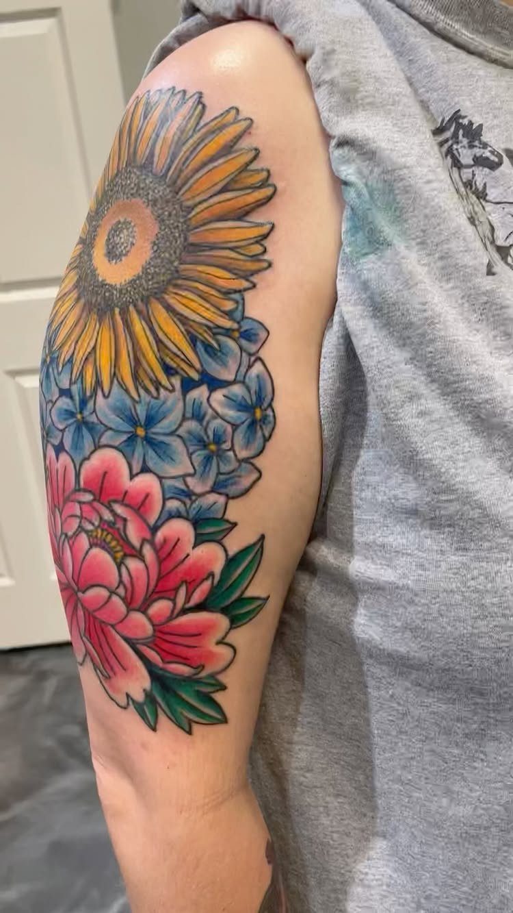 Sunflower, blue flowers, and red peony tattoo on a person's upper arm.