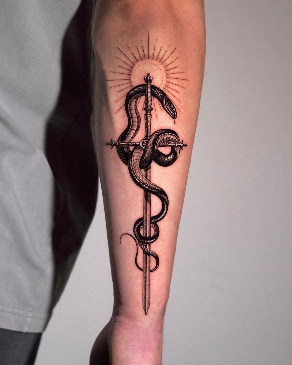 Snake coiled around a sword tattoo on a forearm with a sunburst above.