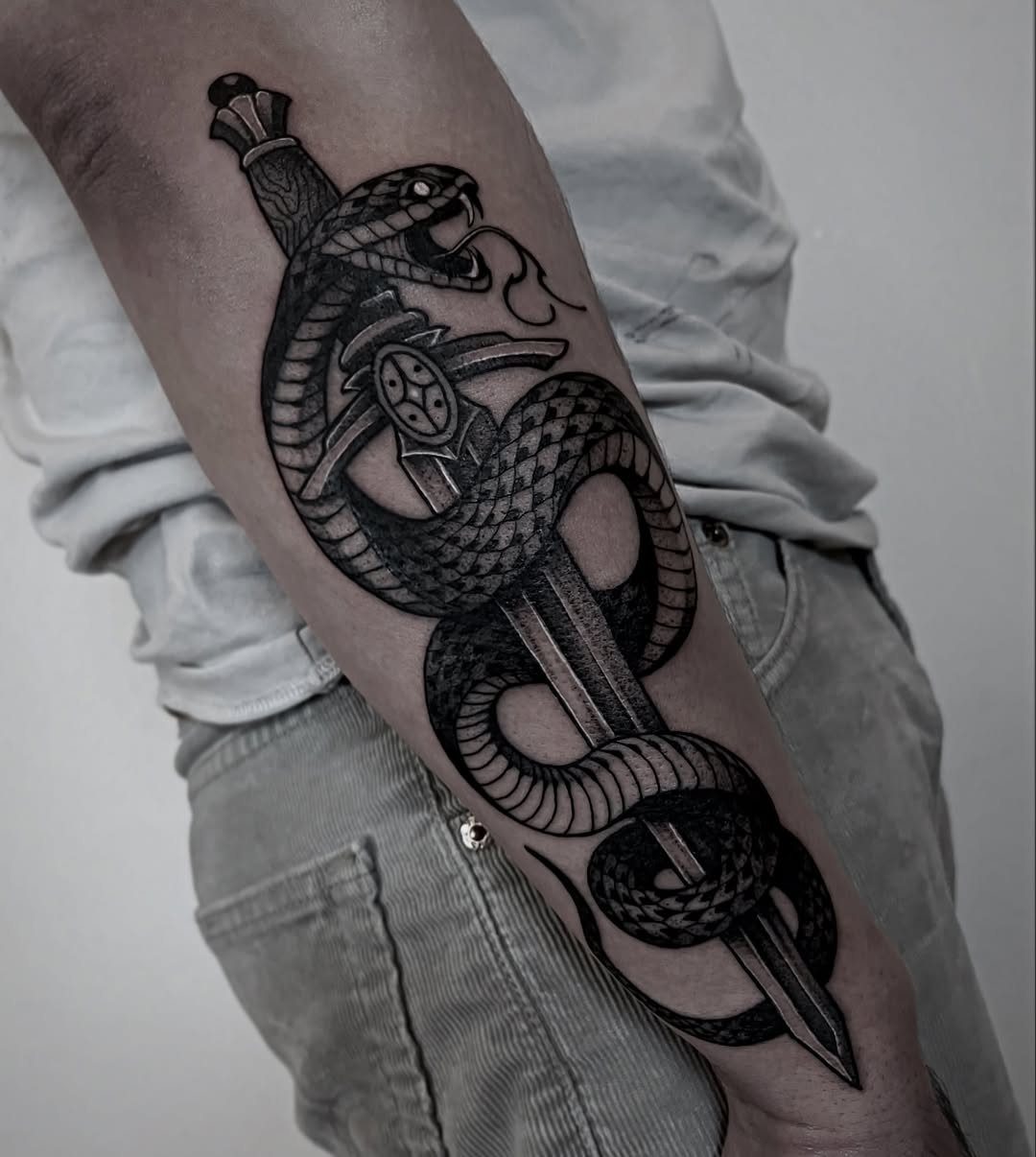 Snake coiled around a dagger tattoo on a person's forearm in black ink.