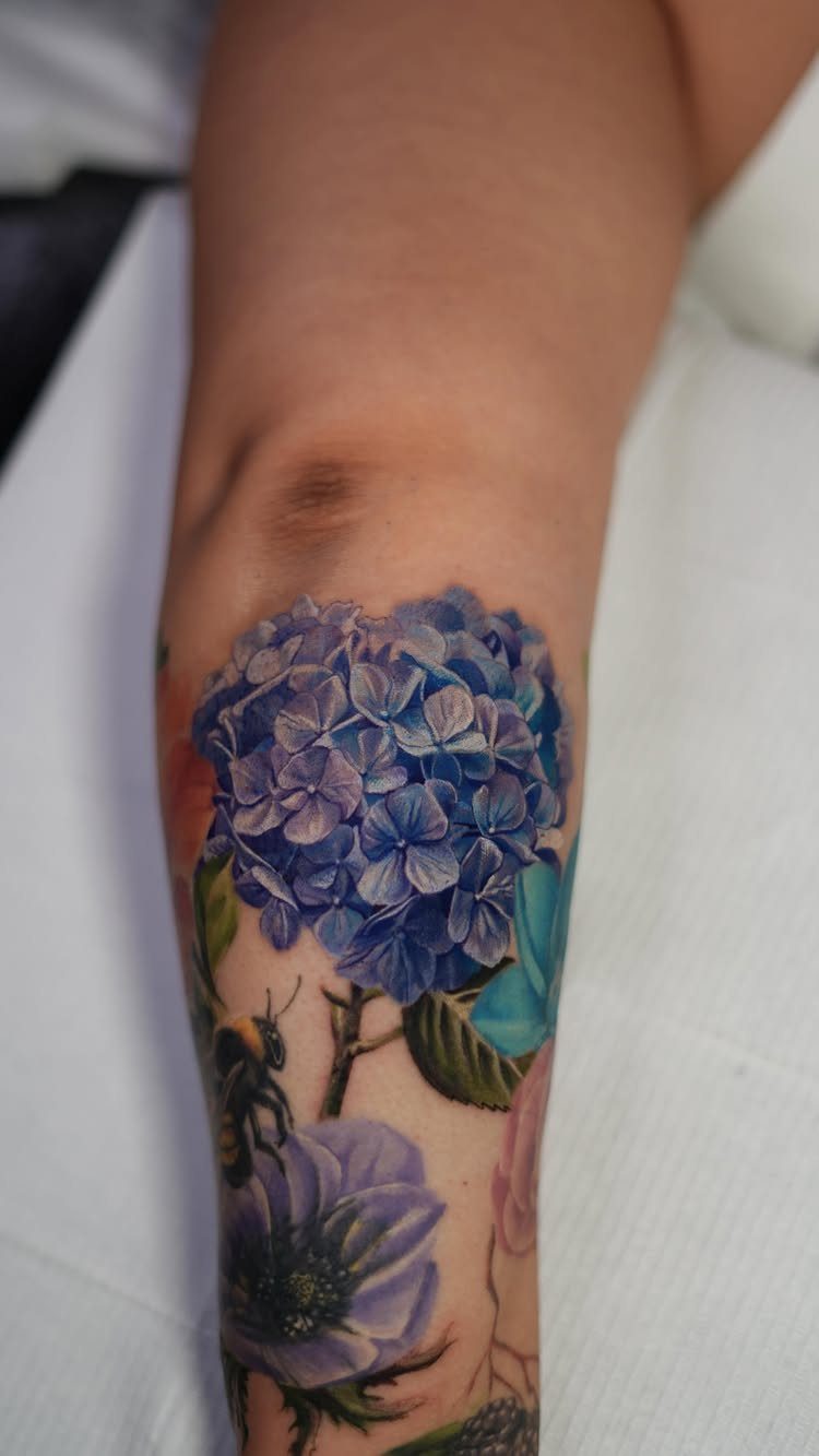 Blue hydrangea cluster tattoo on a forearm with purple flowers and green leaves