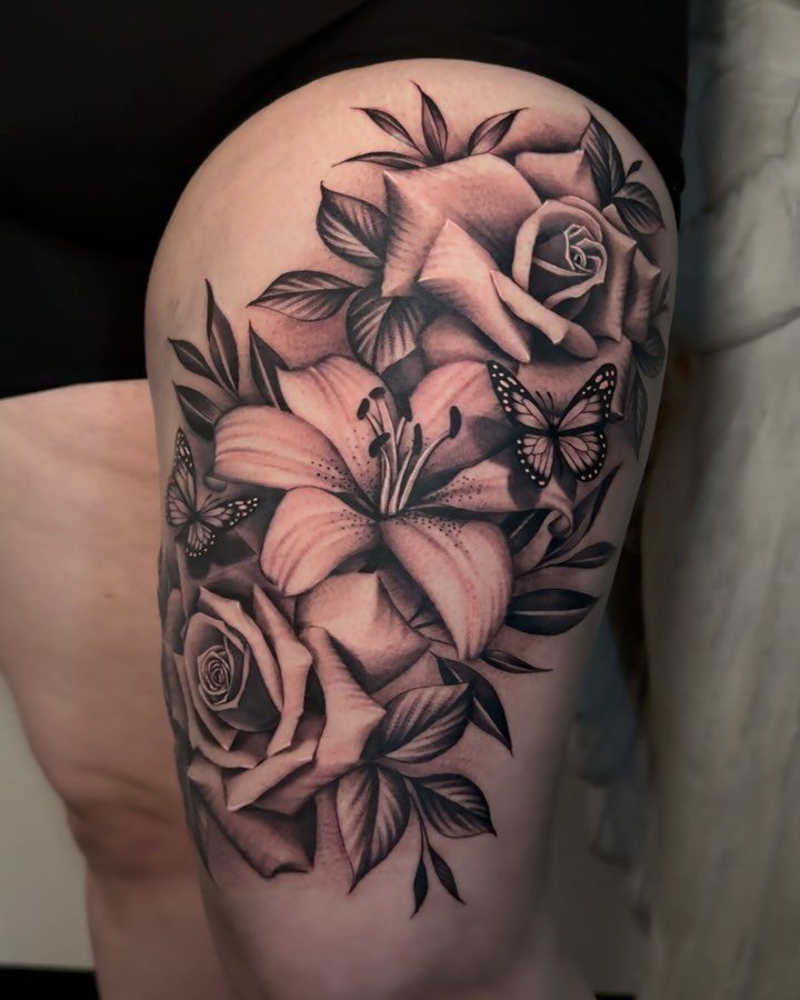 Tattoo on a thigh featuring roses, a lily, and butterflies in black and grey.