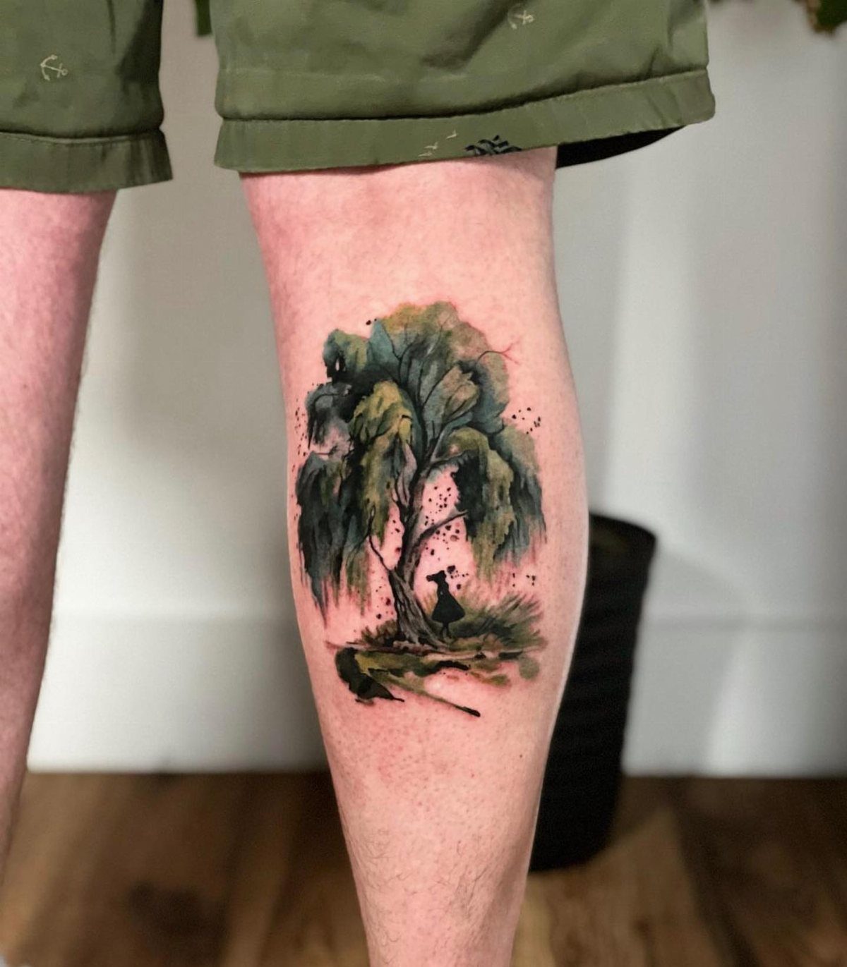 Watercolor weeping willow tree tattoo on a calf with a small silhouette figure.